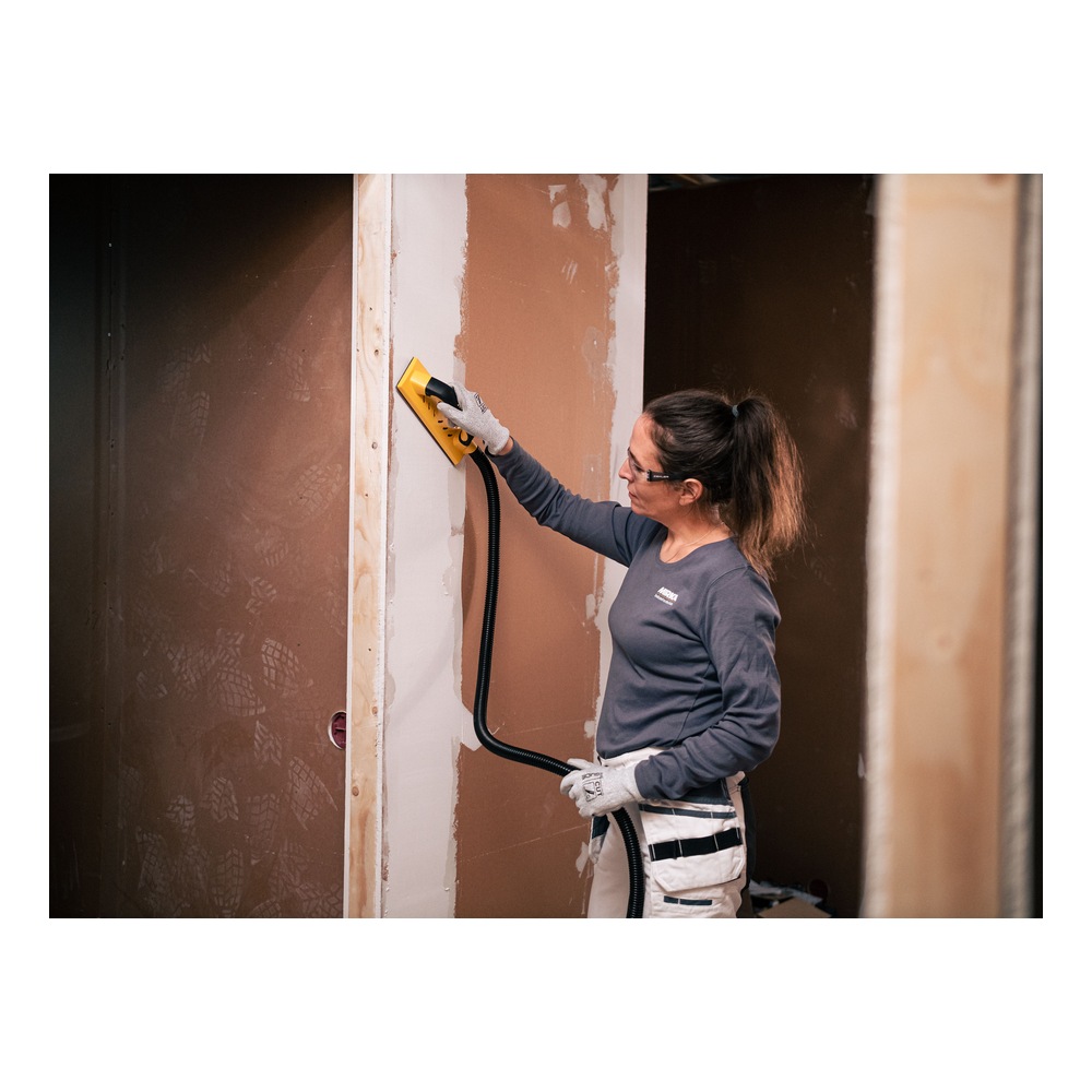 A woman uses a yellow and black hand sander with a suction hose, against