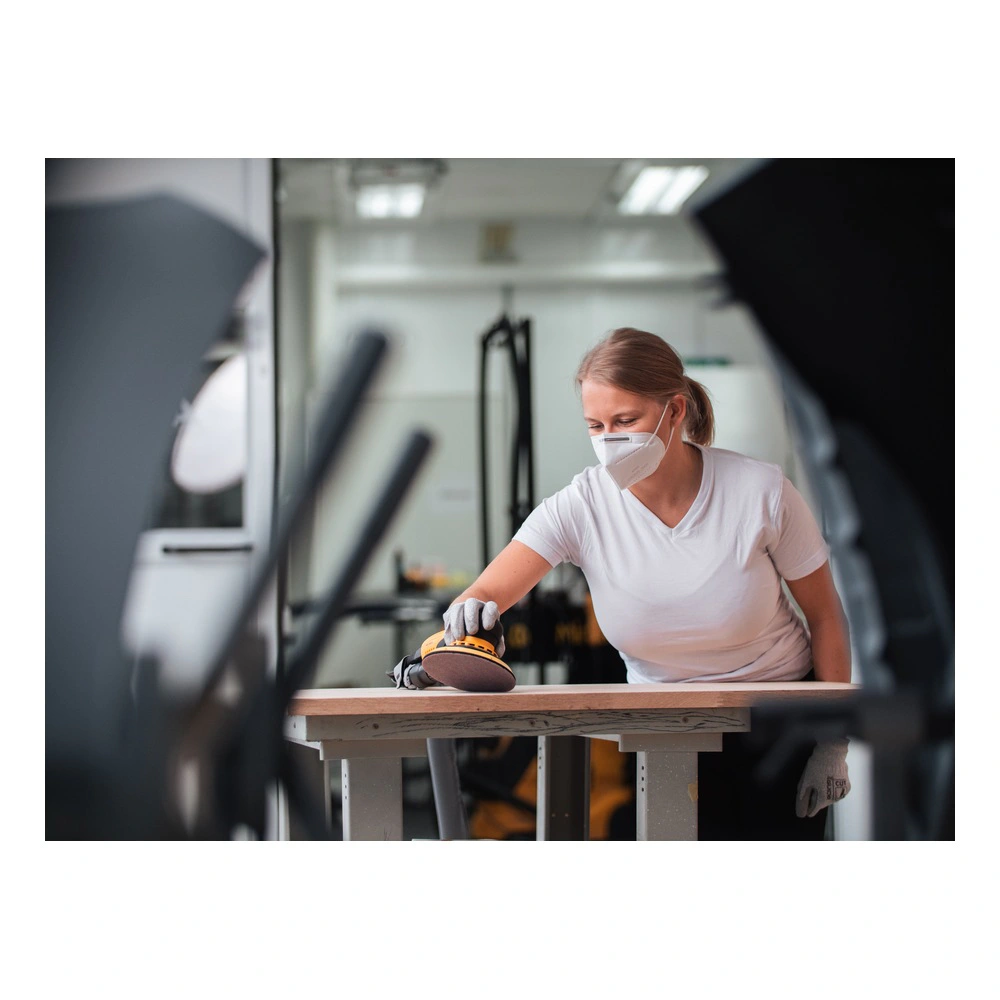 A woman wearing a protective mask uses a handheld sander on a wooden board