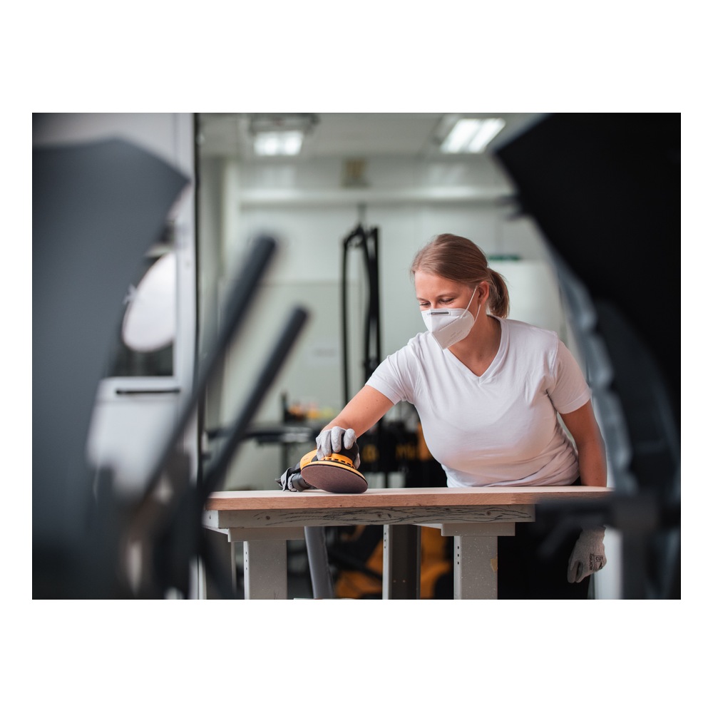 A person wearing a mask sands a wood board with a hand sander, seen through