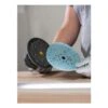 Gloved hands hold two perforated round sanding pads, one black and one blue