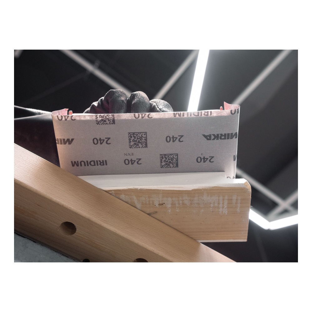 A gloved hand holds a sheet of sanding paper over a wooden piece, with Mirka